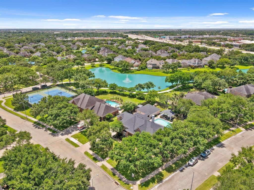 This aerial photo showcases a spacious property with a well-maintained house, a large driveway, and a private pool. The home is surrounded by lush greenery and situated in a quiet neighborhood with nearby homes also featuring pools and landscaped yards.