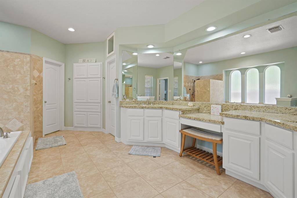 This spacious bathroom features elegant granite countertops, dual sinks, ample cabinetry, and a large vanity area. It includes a walk-in shower and a soaking tub, all set on tasteful tiled flooring. Soft lighting and a soothing color palette create a serene atmosphere.