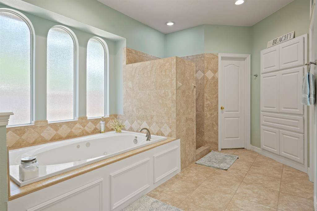 This bathroom features a spacious layout with a large soaking tub, tiled walk-in shower, and ample natural light from three arched windows. Neutral tones and elegant cabinetry enhance the serene atmosphere.