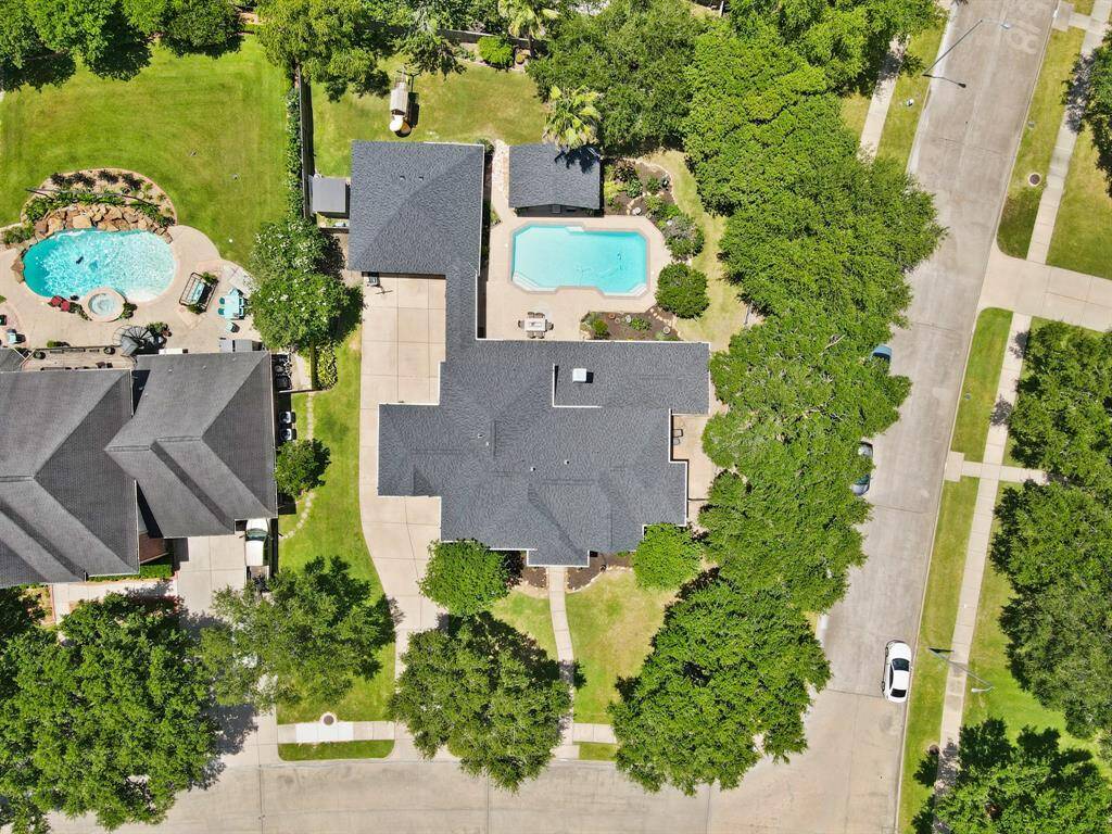 This aerial photo showcases a spacious property with a large house, a swimming pool, and a well-maintained yard surrounded by trees. The home is situated on a quiet street with neighboring houses and ample driveway space.