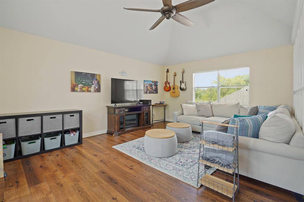 This spacious living room features vaulted ceilings, a large window with natural light, hardwood floors, and a cozy sectional sofa. It's equipped with a ceiling fan, media console, and stylish storage solutions, making it perfect for relaxation and entertainment.
