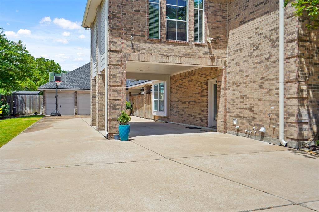 This photo shows a spacious driveway leading to a brick house with a covered area. There's a detached garage with a basketball hoop, and a small lawn on the side. The exterior is well-maintained and inviting.
