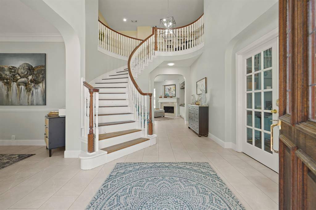 This photo showcases a spacious, elegant entryway featuring a grand, curved staircase with wooden banister, light tile flooring, and soft gray walls. The area is accented with a modern chandelier, decorative artwork, a French door, and leads to a cozy living space with a fireplace.