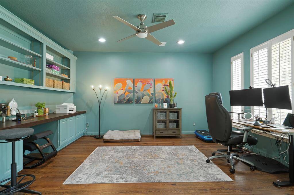 Bright, spacious home office with teal walls, built-in shelving, and a large desk area. Features hardwood floors, ample natural light from large windows, and modern lighting. Ideal for productivity and comfort.