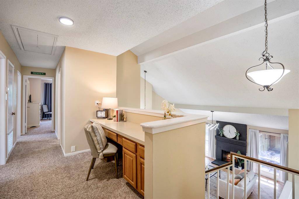 At the top of the stairs, a built-in study nook overlooks the formal living room and fireplace below, creating a bright, open workspace. To the left, the hallway leads to the secondary bedrooms, while just behind you is the private primary suite.