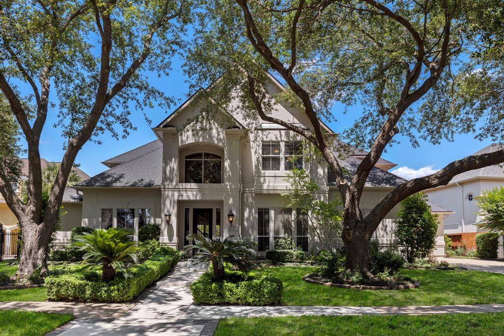 This elegant two-story home features a classic facade with large windows and a welcoming central entrance. It's surrounded by lush landscaping with mature trees and manicured shrubs, offering a serene and attractive curb appeal.