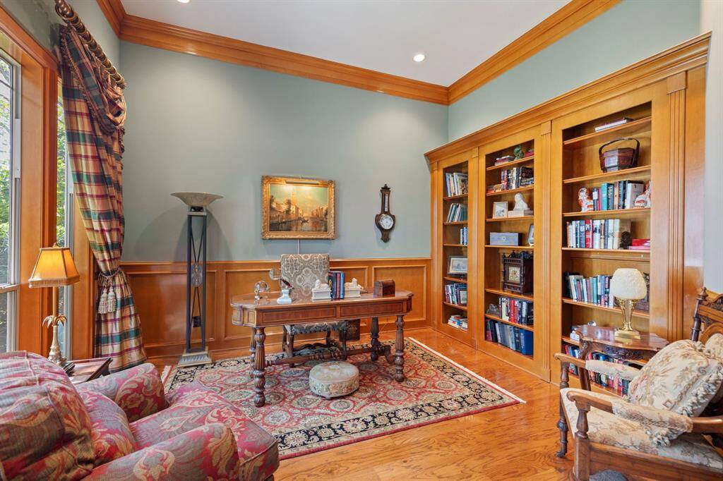 This elegant home office features rich wood paneling and built-in bookshelves, complemented by a large window with luxurious drapes, creating a warm, inviting atmosphere, with a hidden door.