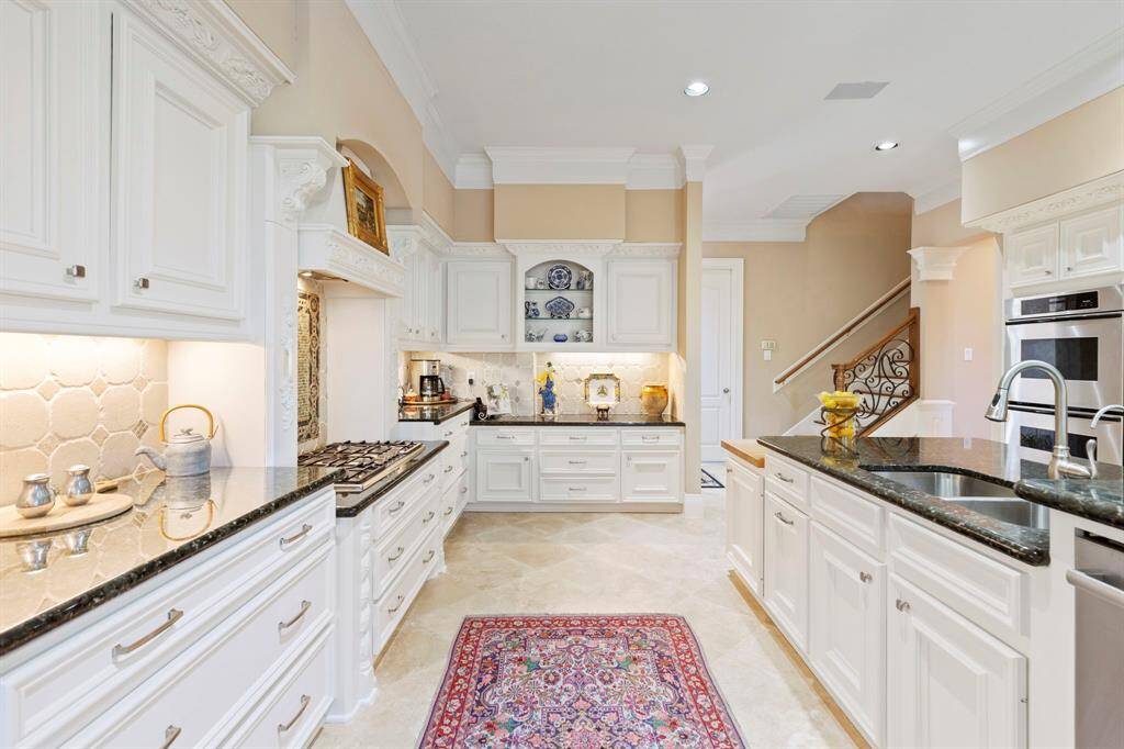 This kitchen features elegant white cabinetry, granite countertops, and a Thermador gas cooktop. It also has Thermador  double ovens, an island with a sink, and a cutting board area. The space is accented with crown molding and recessed lighting, providing a bright and welcoming atmosphere.