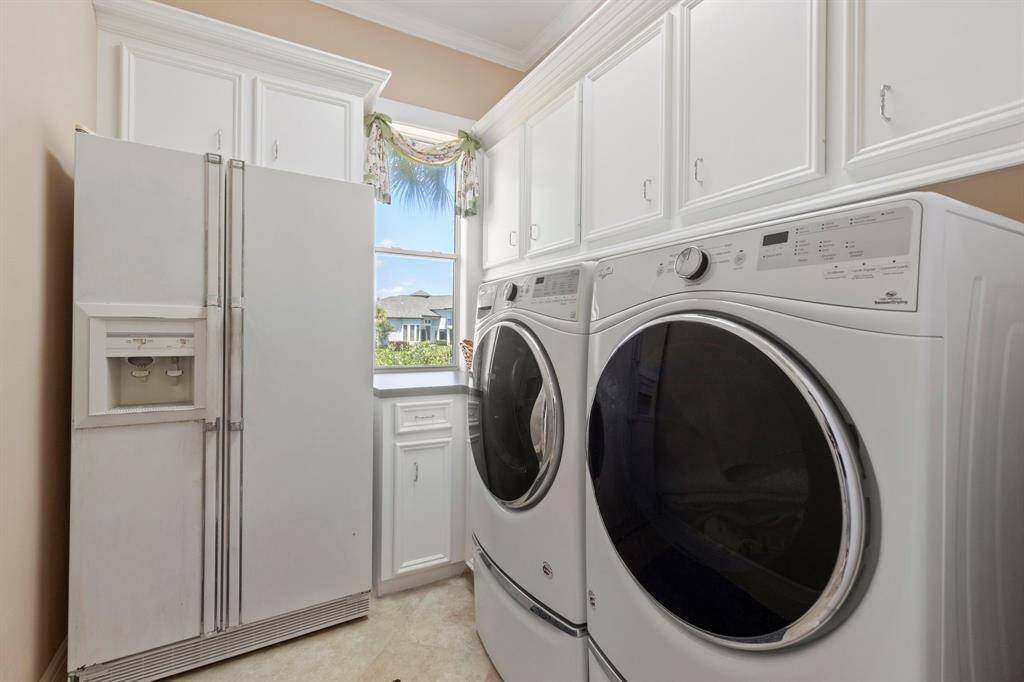 This photo shows a functional laundry room featuring a side-by-side washer and dryer, ample white cabinetry for storage, a large refrigerator with an ice and water dispenser, and a window providing natural light. All appliances stay.