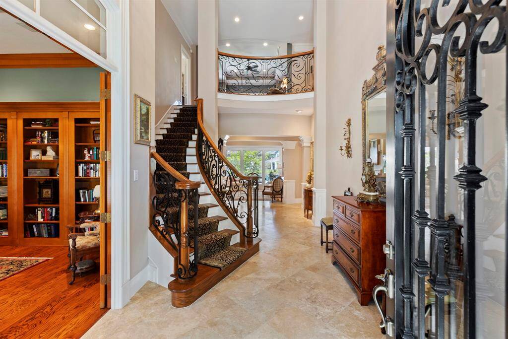 This elegant entryway features a grand staircase with ornate ironwork, a high ceiling, and polished floors. To the left is a cozy library with built-in wooden bookshelves. A decorative mirror and classic furniture enhance the sophisticated atmosphere.