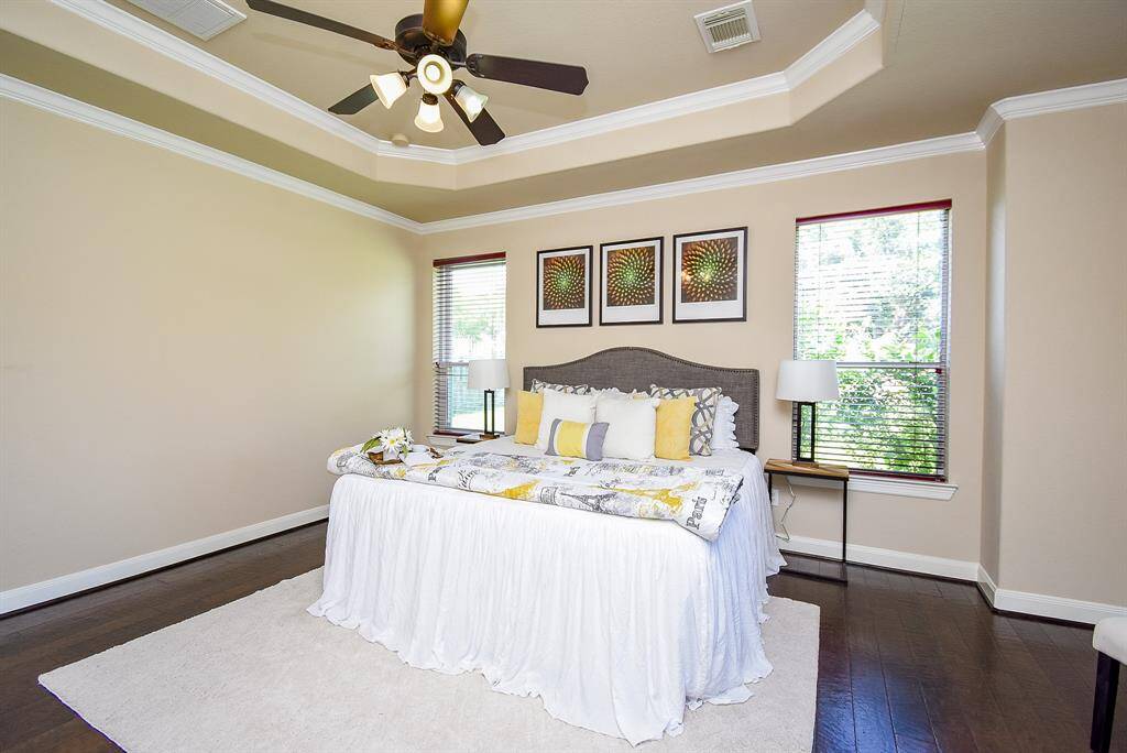 Dark wood floors and soft architectural curves set the tone in the primary bedroom. A tray ceiling and crown molding add depth, while multiple windows ensure a light, tranquil ambiance throughout the day.