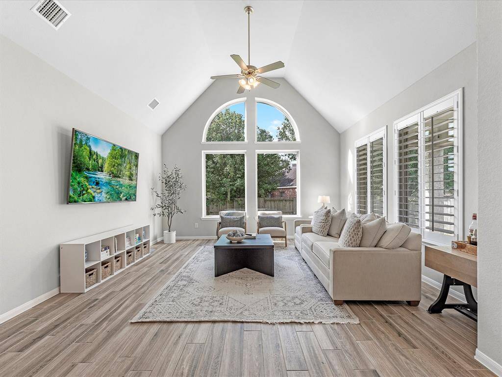 This spacious living room features high vaulted ceilings and abundant natural light thanks to large windows with a scenic view. It includes elegant wood flooring, a cozy neutral-toned sofa, and a mounted TV, offering a comfortable and modern living space.