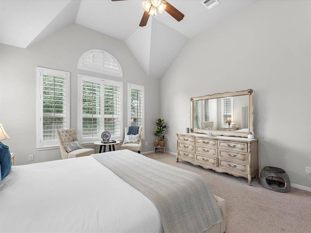 Bright and spacious bedroom featuring high ceilings and large windows with shutters. It includes a cozy seating area, a ceiling fan, and an elegant dresser with a mirror, offering a comfortable and stylish retreat.