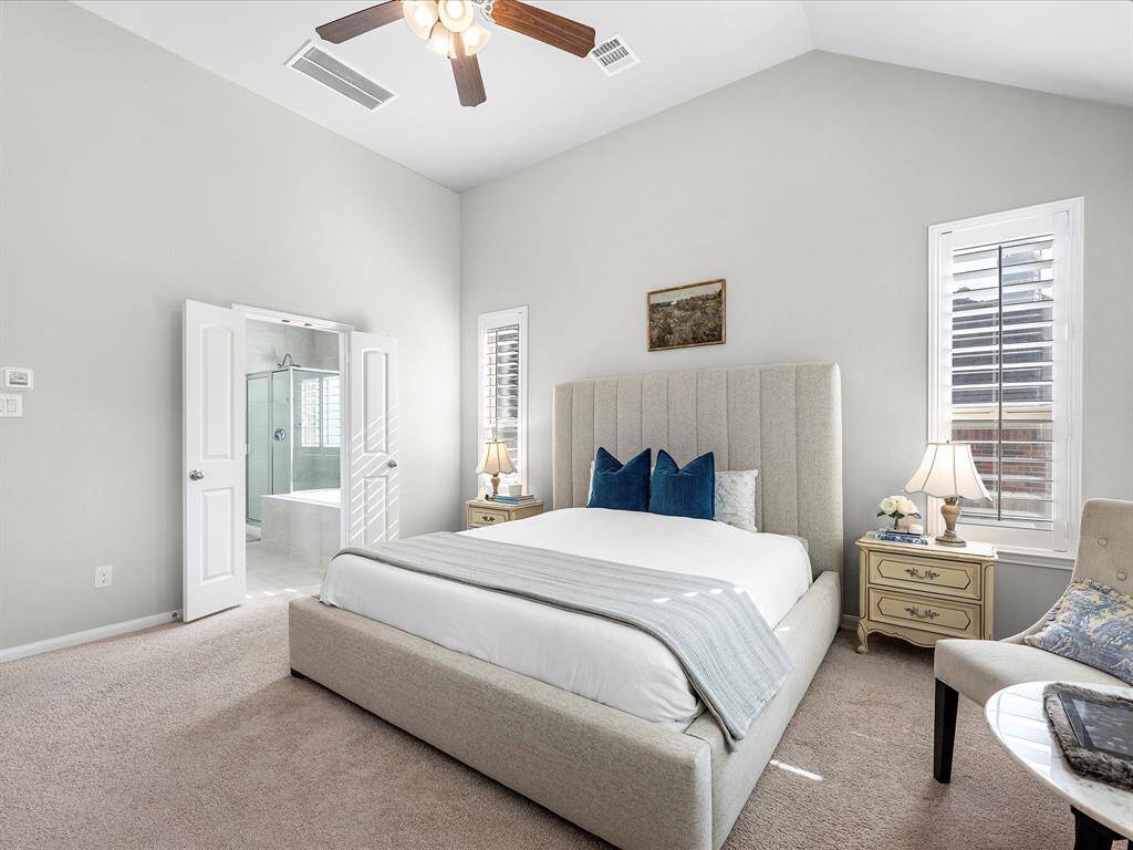 This spacious bedroom features a high ceiling, a comfortable upholstered bed, and abundant natural light from large windows with shutters. It includes an en-suite bathroom with a modern glass shower and tub, adding convenience and luxury.