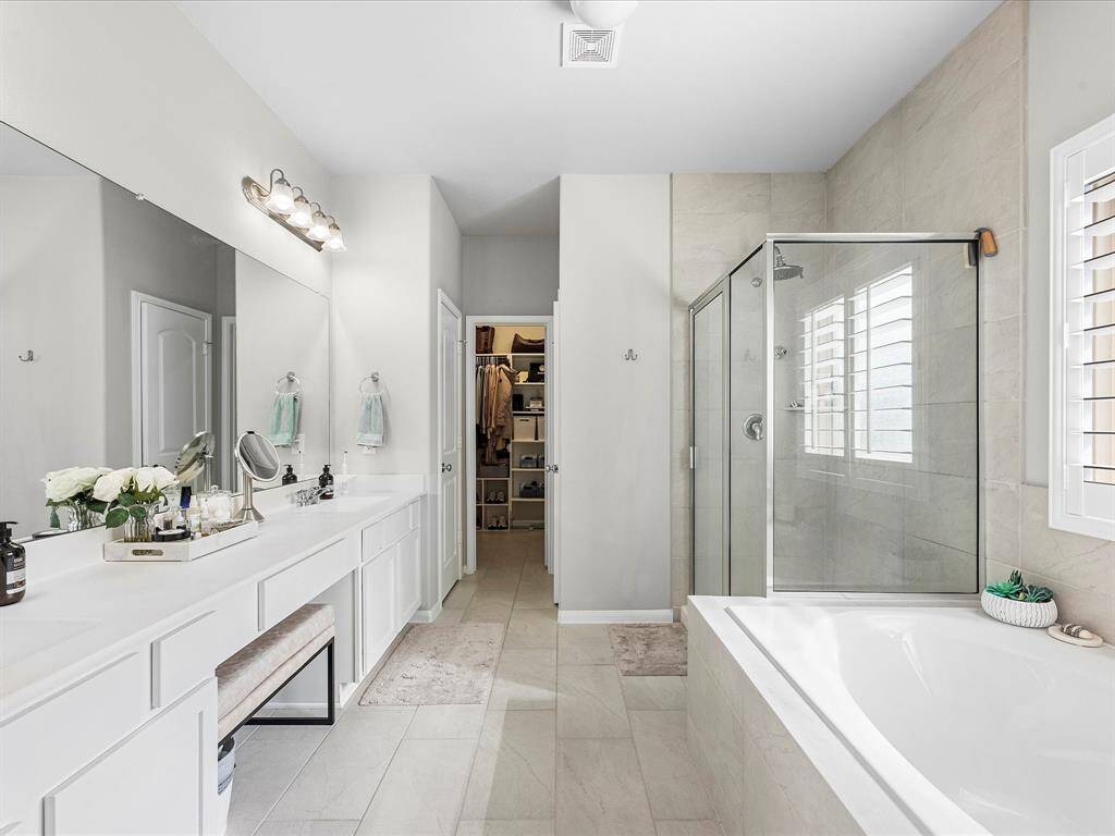 This spacious bathroom features a large double vanity with ample storage, a glass-enclosed shower, and a separate soaking tub. It offers a clean, modern design with neutral tones and plenty of natural light from a window with shutters.