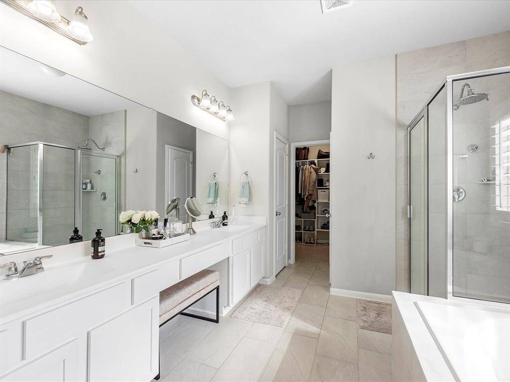 This spacious bathroom features a double vanity with ample counter space, a glass-enclosed shower, and a separate bathtub. It includes modern lighting, neutral tiles, and an adjacent walk-in closet, creating a luxurious and functional space.