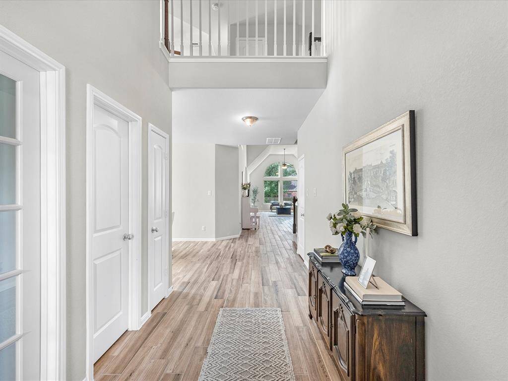This photo showcases a bright, open hallway with wood flooring, leading to a living area. It features a high ceiling with a second-floor overlook, a decorative console table, and natural light from large windows in the background.