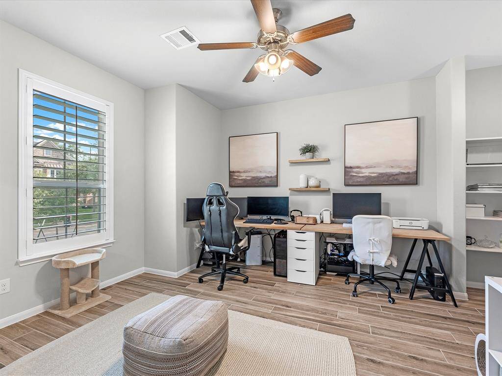This photo showcases a bright, well-organized home office with modern decor. It features dual workstations, a ceiling fan, wood-look flooring, and a large window with shutters for natural light. The space is stylish and functional, perfect for remote work.