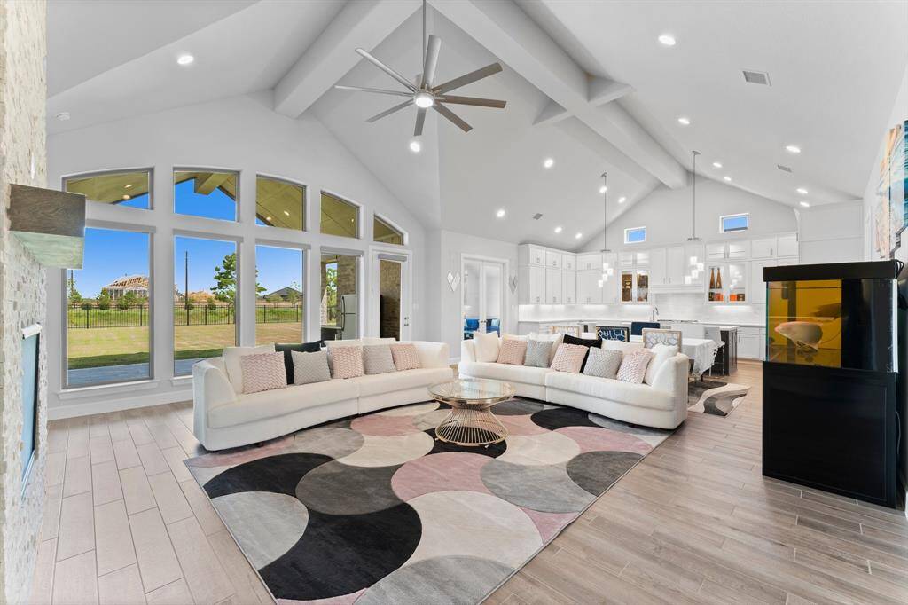 This is a spacious, modern living room with high vaulted ceilings, abundant natural light from large windows,  open floor plan that extends into a sleek kitchen. The room features a stylish  fireplace and offers views of the outdoor space.