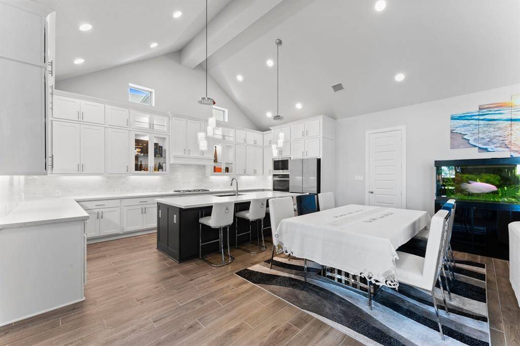 This photo shows a modern, open-concept kitchen and dining area with white cabinetry, stainless steel appliances, a central island with seating, and a spacious dining  area, all complemented by high ceilings and ample recessed lighting.