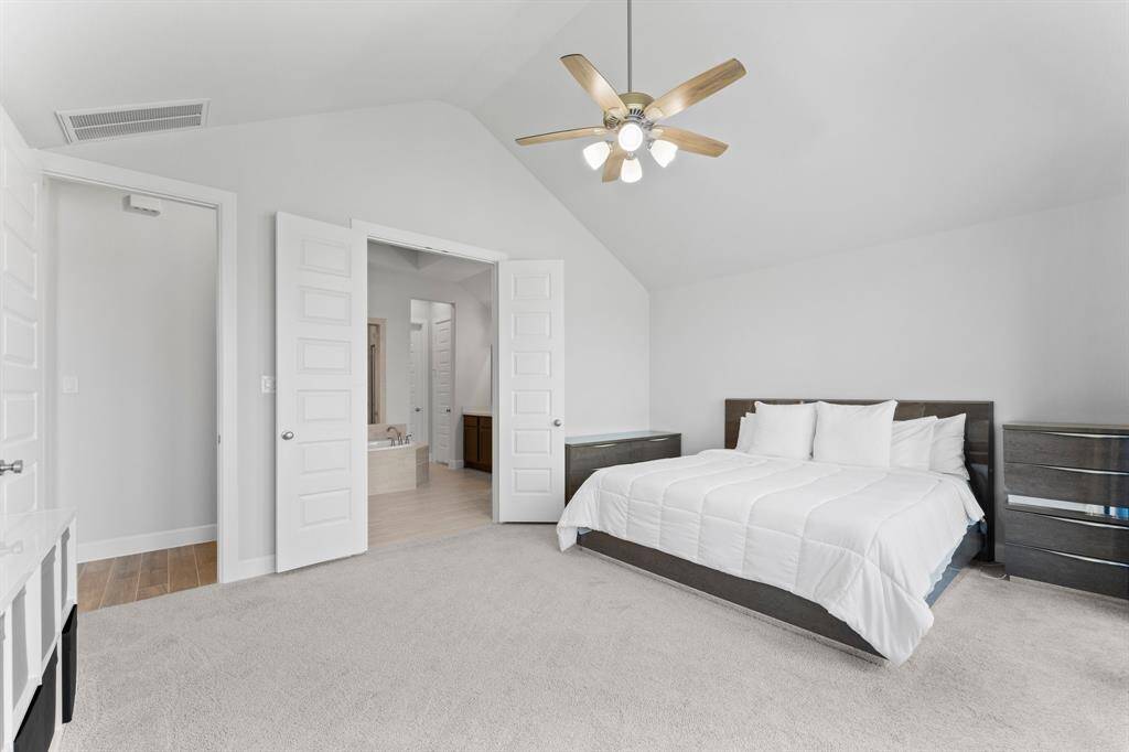 Another view of this master retreat  with vaulted ceiling, plush carpeting, and a neutral color scheme. There's a modern ceiling fan, the room features direct access to an en suite bathroom, offering convenience and privacy.