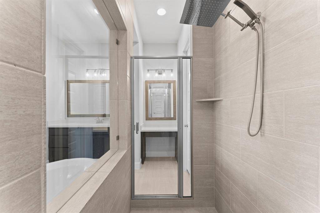 bathroom featuring a large walk-in shower with a rainfall shower head, neutral-toned tile walls, a sleek glass door, and a built-in niche for toiletries. A bathtub and vanity with a mirror are partially visible, suggesting a well-appointed, cohesive design.