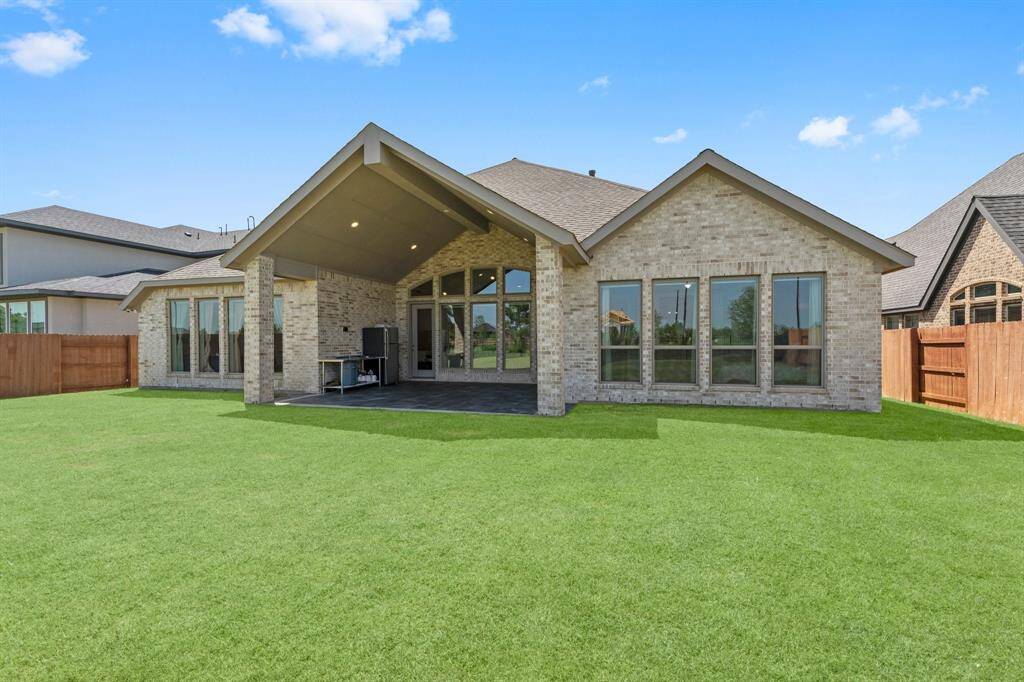 large covered patio area, brick exterior, and expansive windows. The well-maintained lawn is enclosed by a wooden fence, providing privacy and a safe space for outdoor activities.