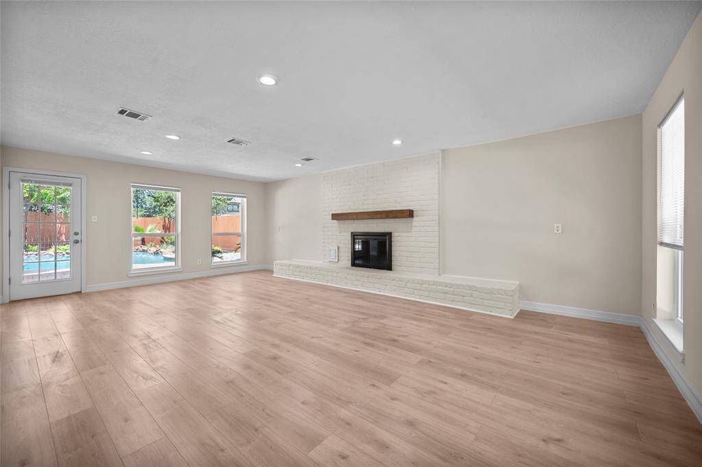 Bright and spacious living room featuring a white brick fireplace, vinyl flooring and large windows with views of the backyard pool.