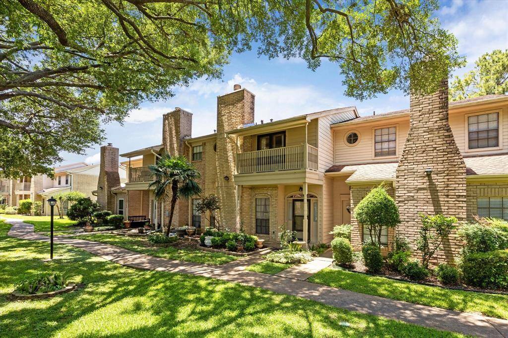 This photo shows a charming two-story townhouse with brick exteriors and a balcony. It features lush landscaping, a paved walkway, and mature trees, offering a serene and inviting atmosphere.