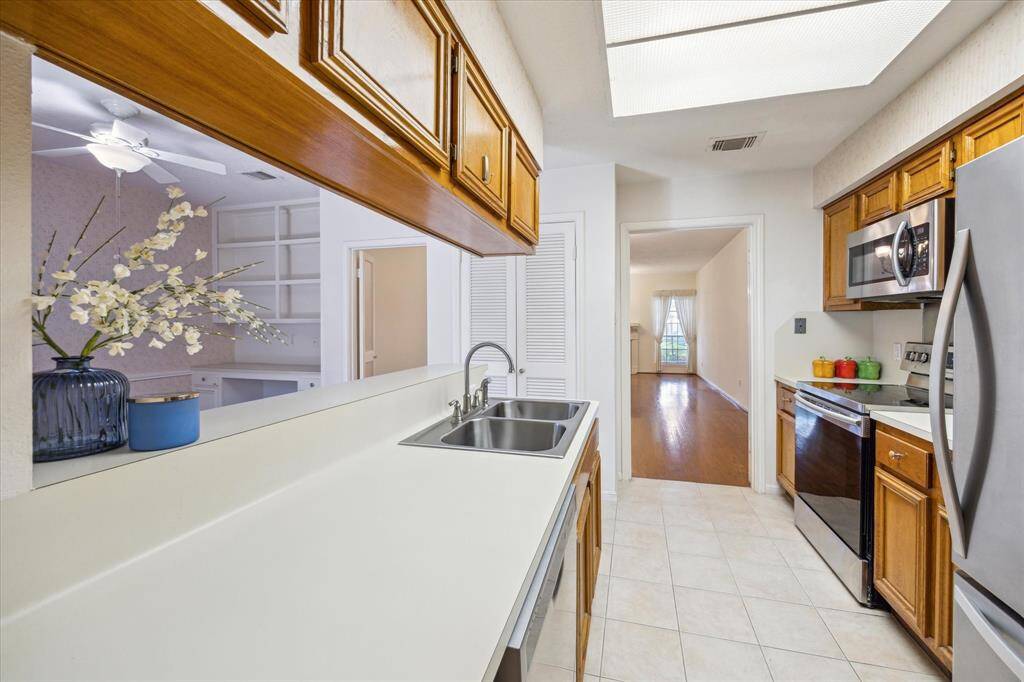 This kitchen features modern stainless steel appliances, wooden cabinetry, and a double sink. It opens to a bright living area with a hardwood floor and a large window, creating a seamless flow throughout the home.