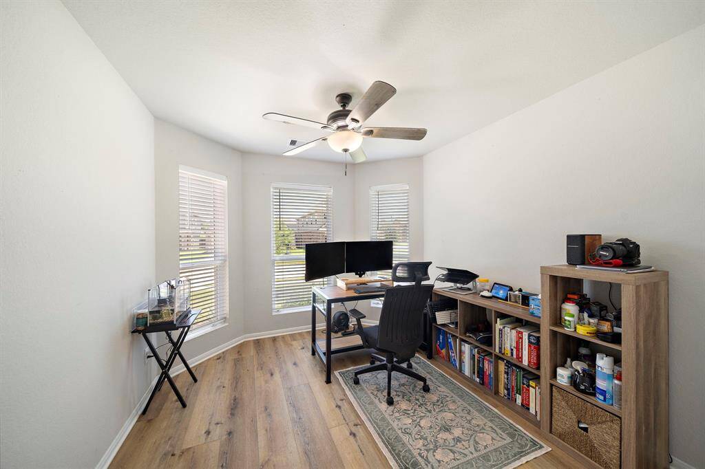 Bright home office with natural light from multiple windows, featuring a ceiling fan, high-end flooring, and ample space for a desk and shelving.