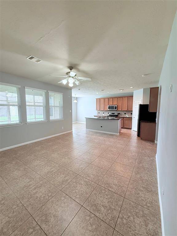 This spacious open-plan area features a kitchen with a central island, modern appliances, and ample cabinetry. The adjoining living space has large windows for natural light, tiled flooring, and a ceiling fan, creating a bright and welcoming environment.