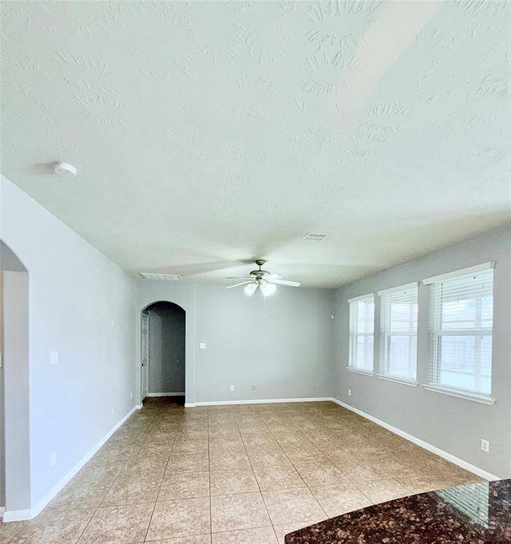 Spacious living room with elegant tile flooring, abundant natural light from large windows, and a ceiling fan for comfort. Perfect for relaxing or entertaining.