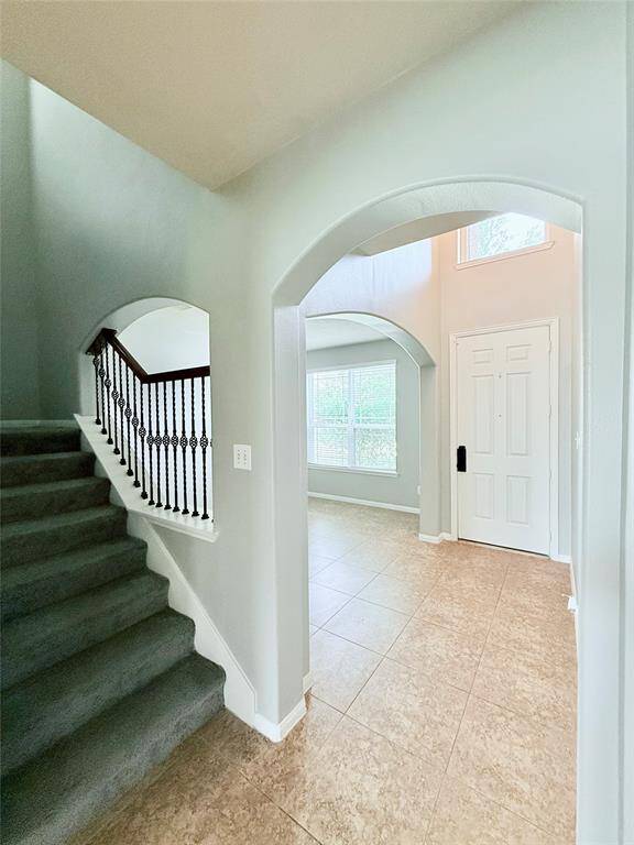 A bright entryway with a tiled floor and high ceilings. It features an arched doorway, a staircase with a wrought iron railing, and a window allowing natural light to fill the space.
