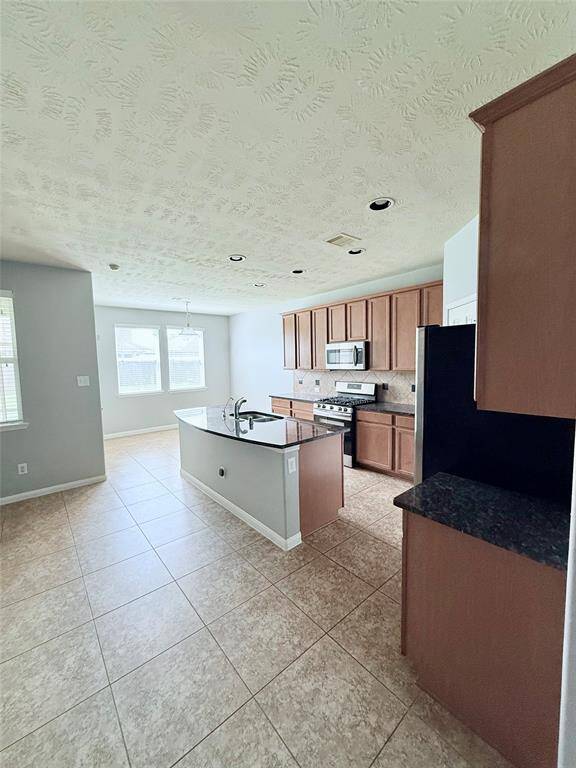 Modern kitchen with ample natural light, featuring tiled floors, a central island with a sink, stainless steel appliances, and wooden cabinetry.