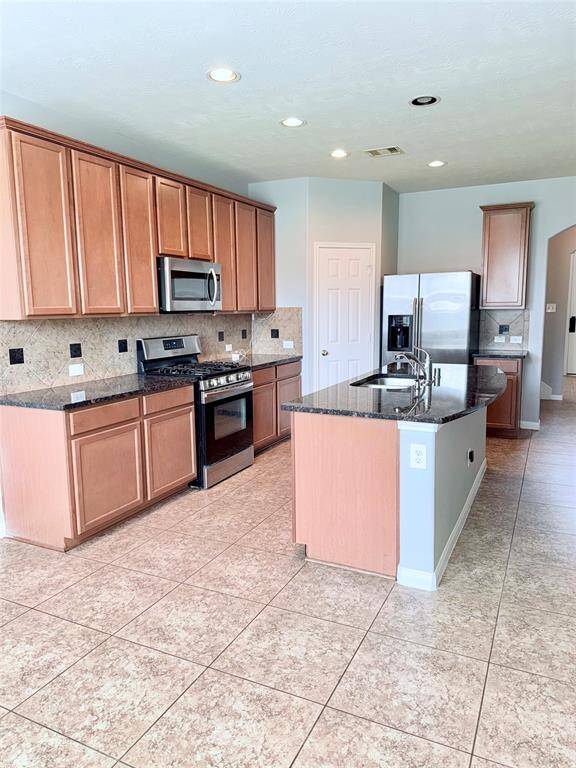 Spacious kitchen with modern stainless steel appliances, granite countertops, and ample wooden cabinetry. Features a central island with a sink, tiled floors, and bright recessed lighting.