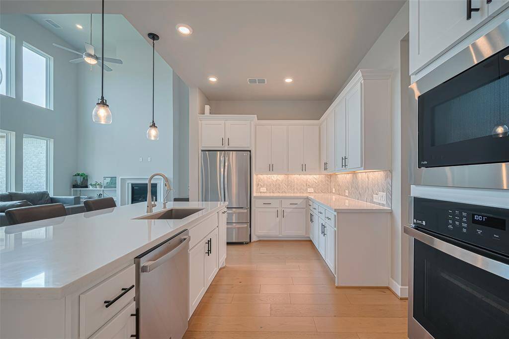 This modern kitchen features sleek white cabinetry, stainless steel appliances, and a spacious island with a built-in sink. Pendant lighting adds elegance, while the open layout provides a seamless flow to the living area with high ceilings and large windows.