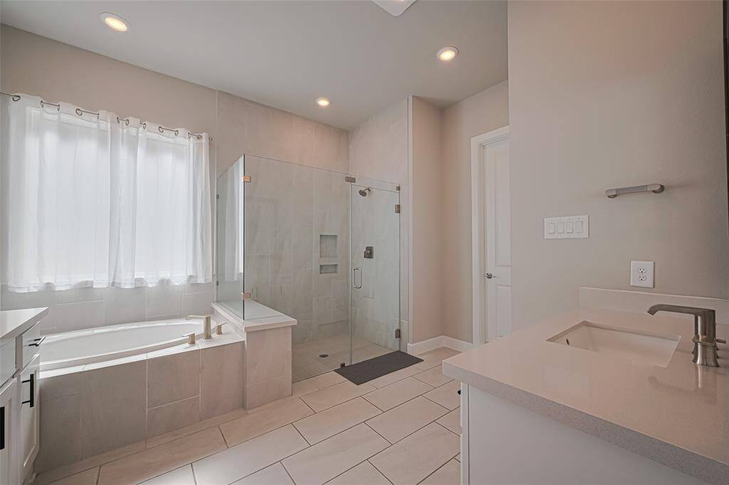 Primary bathroom features a spacious layout with a glass-enclosed shower, a large soaking tub, and a sleek vanity. Natural light fills the room through a window with sheer curtains, creating a bright and inviting space.