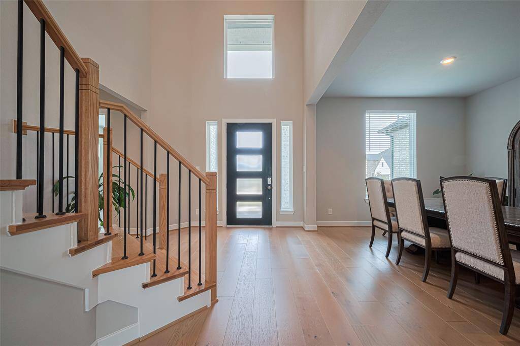 A bright, open entryway with high ceilings w/ modern design. A stylish staircase with wood and iron railing, a contemporary front door with glass panels, and an adjacent dining area with large windows, providing ample natural light.
