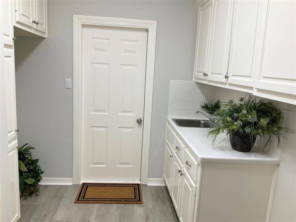 Bright utility room with white cabinetry, a sink, and new flooring. Lots of cabinetry in the utility room with a sink and room for another refrigerator in the space on the left.  The door leads to a spotless, oversized garage with epoxy paint flooring.The home or streets have never flooded in this community.