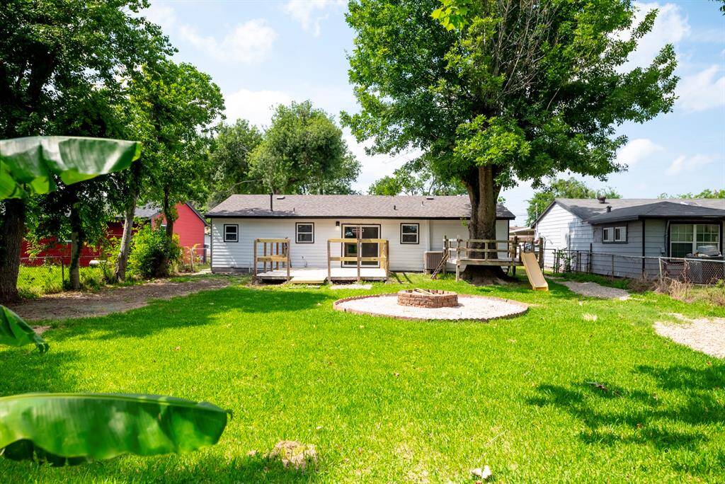 Charming and oversized backyard with lush green grass, mature trees, and a cozy fire pit area. The house features a small deck and a slide, perfect for family activities and outdoor gatherings.