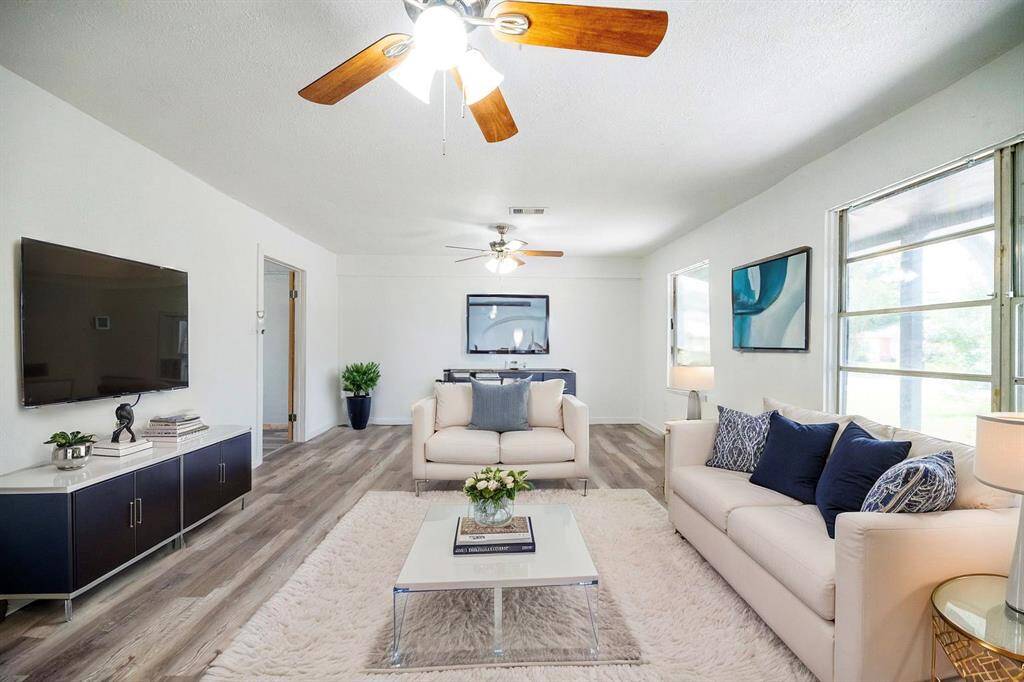 Bright and airy living room with modern decor, featuring light wood flooring, cozy sofas, a large wall-mounted TV, and ample natural light from large windows. Ceiling fans and stylish accents create a welcoming atmosphere. This picture is virtually staged