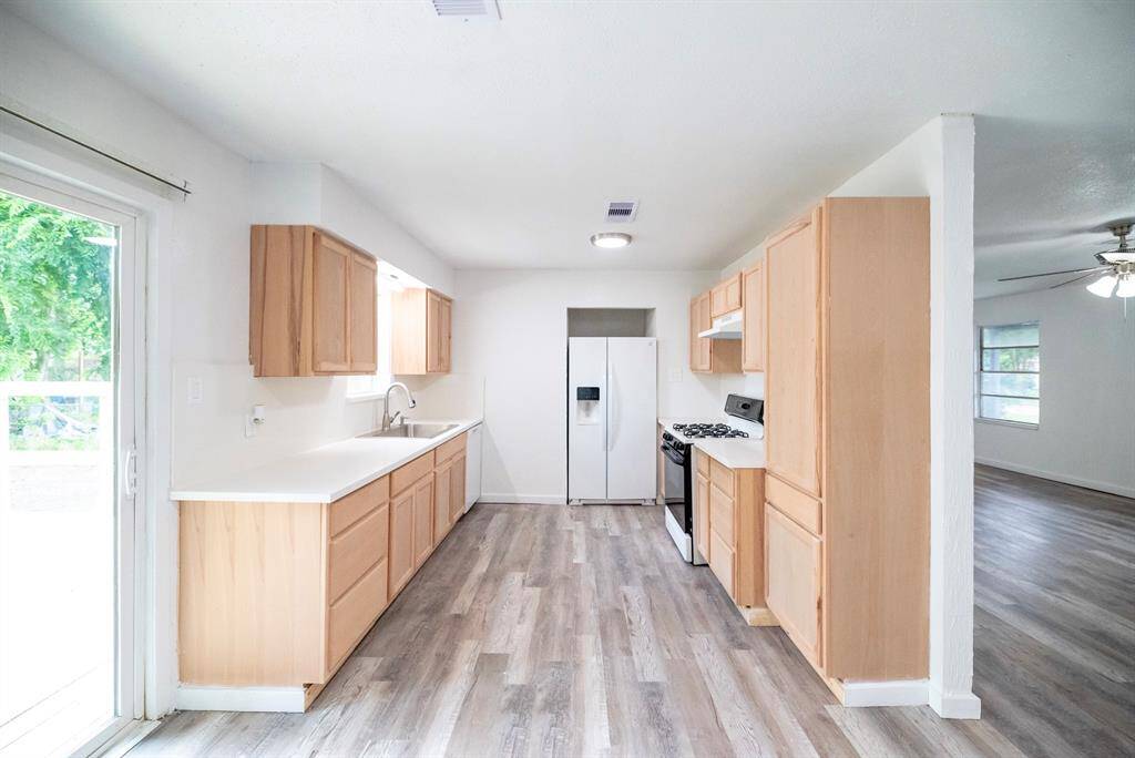 This photo showcases a bright, open kitchen with light wood cabinets, white countertops, and stainless steel appliances. It features a gas stove, double-door refrigerator, and ample natural light from a large sliding glass door and window, leading to a spacious adjacent living area with wood-look flooring.