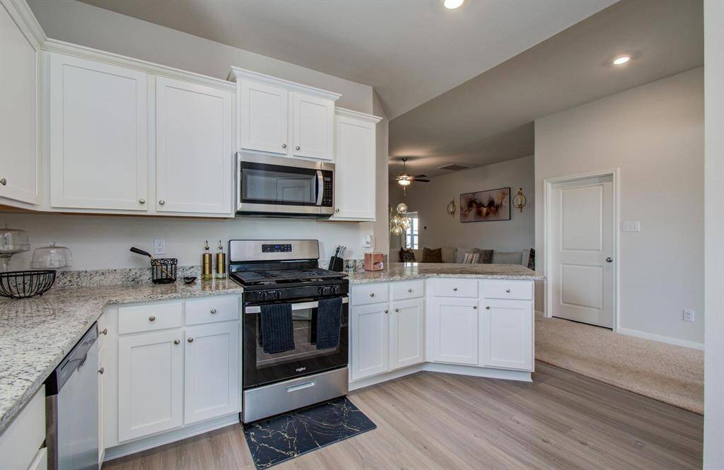 This kitchen features modern white cabinetry, granite countertops, and stainless steel appliances. It opens into a cozy living area with a ceiling fan and neutral decor, providing a seamless and inviting space.