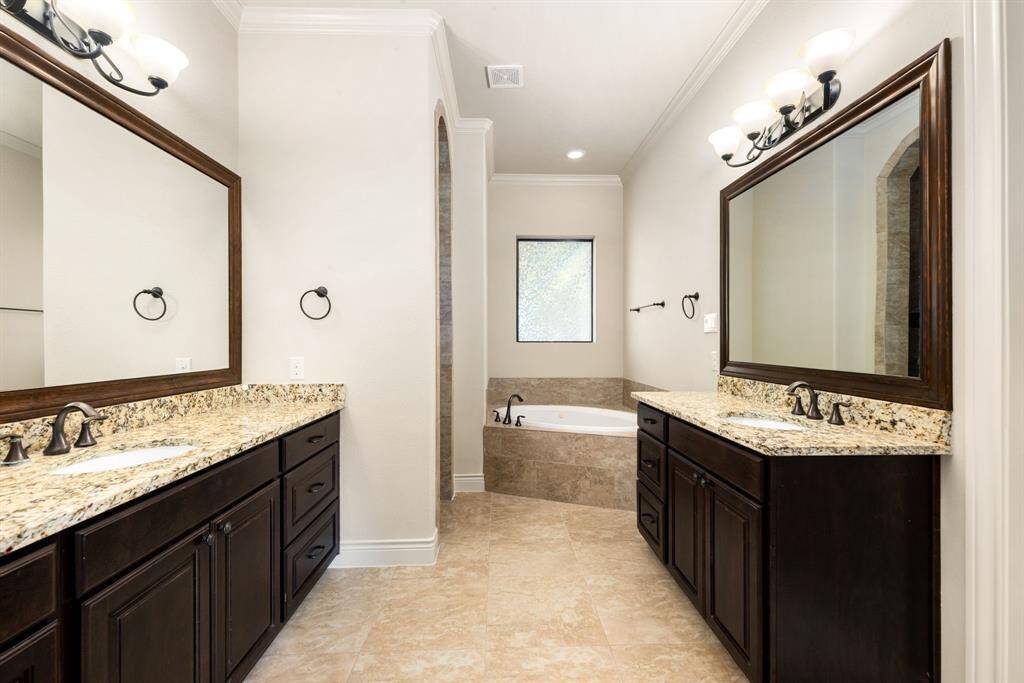 This spacious primary ensuite bathroom features dual granite-topped vanities with dark wood cabinetry, a large soaking tub with separate shower, and elegant light fixtures, creating a luxurious and functional space.