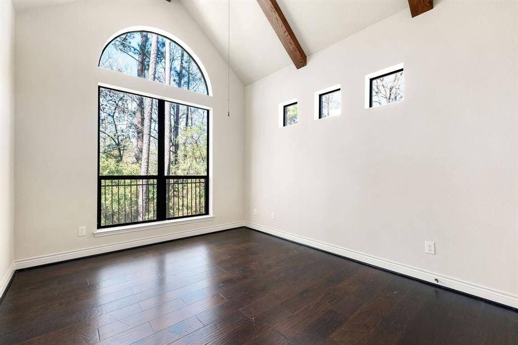 This secondary bedroom features high ceilings with exposed wooden beams and rich dark hardwood floors. It includes a large arched window offering scenic views of trees, allowing natural light to fill the space.