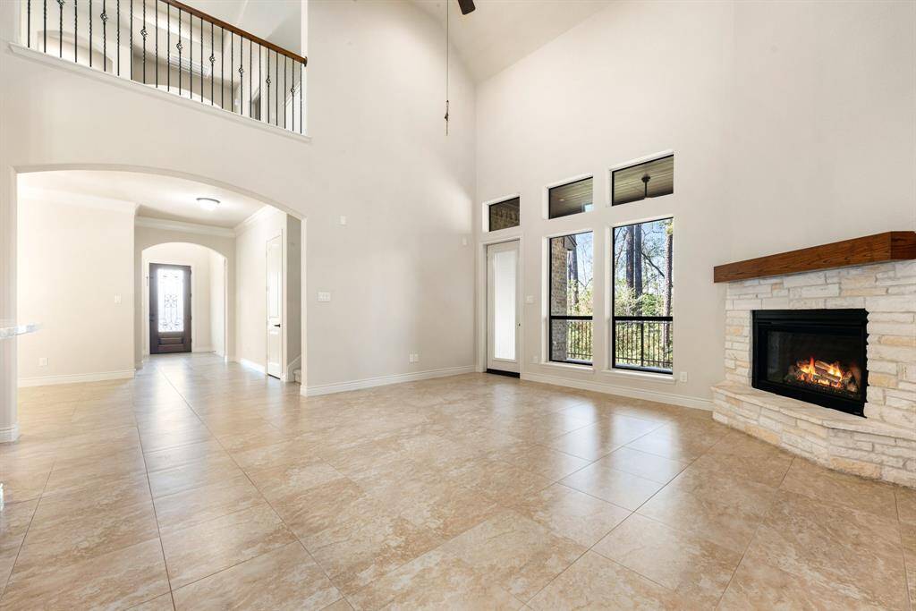 Bright and spacious living area with high ceilings, large windows, and a cozy stone fireplace. The room features tile flooring and opens to a hallway leading to the front door. Ideal for modern living with plenty of natural light.