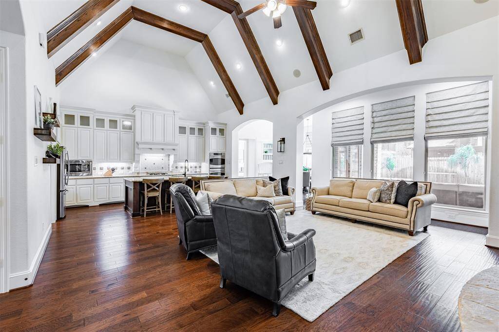 This spacious living area features high vaulted ceilings with exposed wooden beams, a bright open floor plan, and a modern kitchen with white cabinetry. The room is accented by hardwood floors and large windows that allow natural light to flood the space, creating a warm and inviting atmosphere.