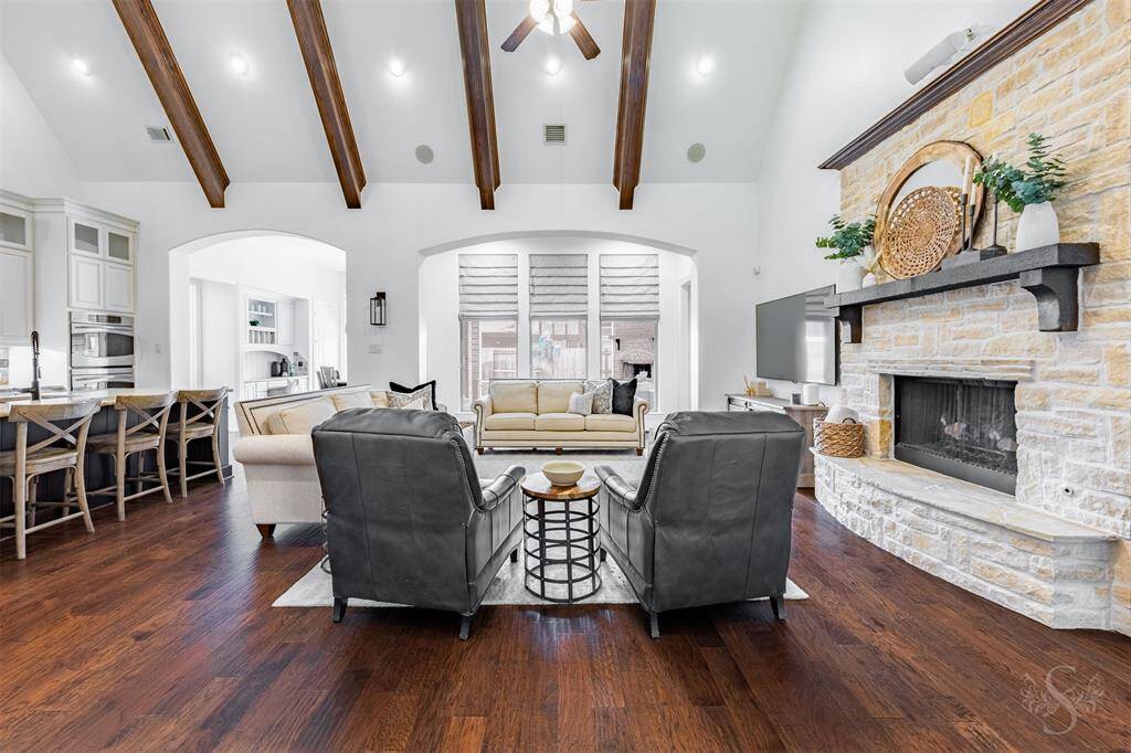 This inviting living space features high ceilings with wooden beams, a cozy stone fireplace, and elegant hardwood floors. The open layout connects seamlessly to a modern kitchen with a breakfast bar, creating a perfect setting for relaxation and entertaining. Natural light pours in through large windows, enhancing the room's warmth and charm.