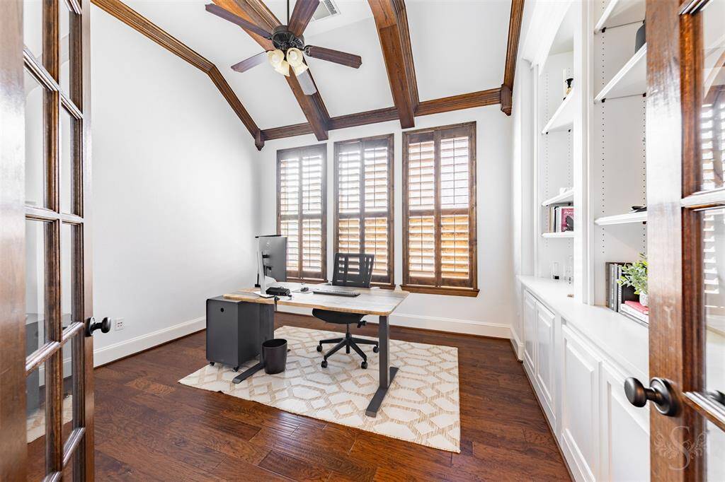 This bright home office features high ceilings with exposed wooden beams and large windows with shutters, allowing plenty of natural light. It includes a ceiling fan, built-in shelving for storage and is finished with hardwood floors. Glass enclosed French doors add privacy to the room when needed.